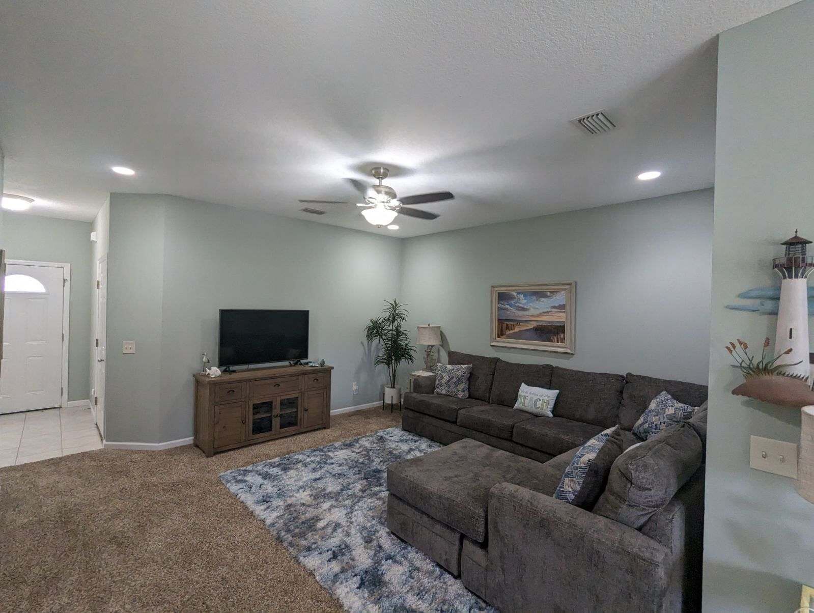 Un salon doté d’un ventilateur de plafond, d’un téléviseur à écran plat posé sur un meuble en bois, d’un canapé gris agrémenté de coussins à motifs et d’un tapis bleu et blanc.