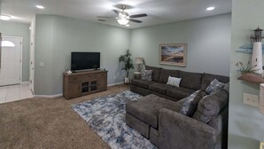 Una sala de estar con un ventilador de techo, un televisor de pantalla plana sobre un mueble de madera, un sofá gris con cojines estampados y una alfombra azul y blanca.