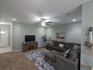 A living room with a ceiling fan, a flat-screen TV on a wooden cabinet, a grey couch with patterned cushions, and a blue and white rug.