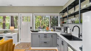 A modern kitchen with grey cabinets, a white countertop, and a black sink. There is a window with a view of trees, and a small table with a lamp and a bowl of fruit. - Private Urban Forest Hideaway in the Heart of Portland with AC (Portland)