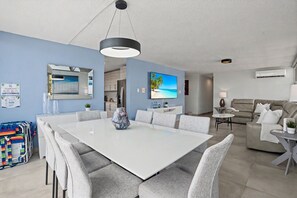 Un salon moderne avec une grande table à manger blanche, des canapés gris, un téléviseur et un tableau sur le thème de la plage.