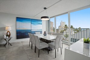 A modern dining area with a large table and chairs, a painting on the wall, and a balcony with a view of buildings and palm trees. - Caribe Aqua Loft  Steps to the Beach  3BR Condo (Isla Verde)