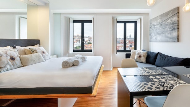 Une chambre d’hôtel moderne dotée d’un grand lit, d’un canapé, d’une table à manger et de deux fenêtres donnant sur la ville.