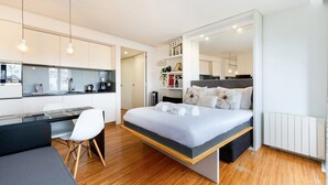 Une chambre d’hôtel compacte comprenant un coin cuisine, un lit avec une literie à motifs floraux, un bureau avec une chaise et un grand miroir.
