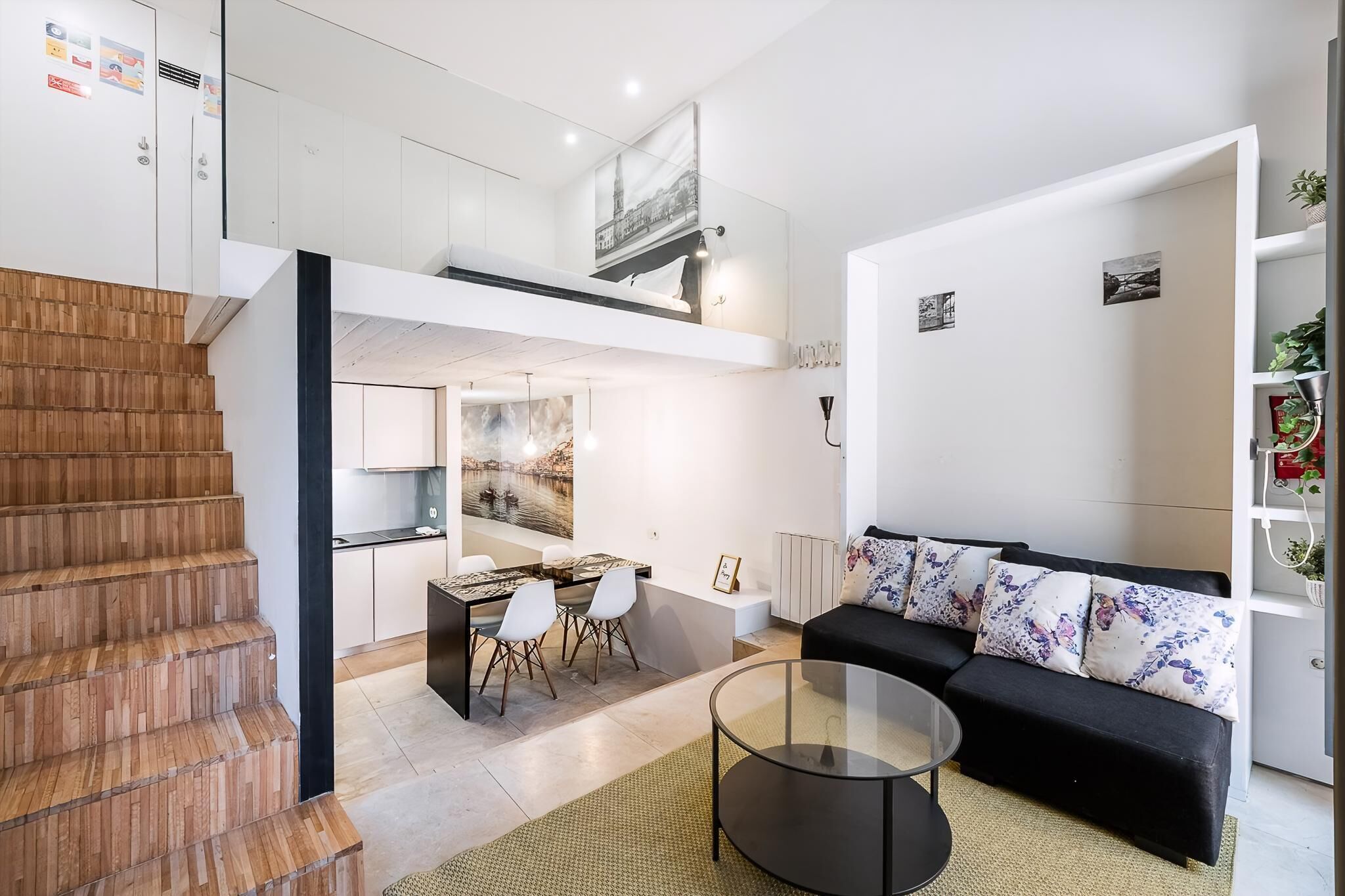 A modern apartment with a staircase, a dining area, a black sofa, and a glass coffee table.
