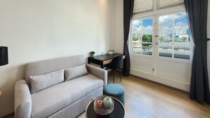 Una sala de estar moderna con un sofá gris, una mesita redonda con un cuenco decorativo, una silla negra y una ventana con cortinas.