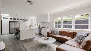 En rummelig opholdsstue med en brun lædersofa, et hvidt sofabord og et køkken med hvide skabe samt et stort vindue.