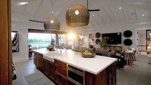 A modern kitchen with a large island, wooden cabinets, and a built-in oven. There is a dining area with a TV and decorative wall art. - 4BR I Kii Hale at Kukui ula Signature Retreat (Poipu)