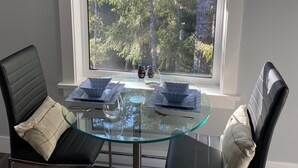 A dining area with a glass table, two black chairs, and a window with a view of trees.