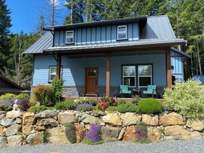 A two-story house with a stone foundation, a front porch with two chairs, and a stone wall with colorful flowers.