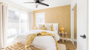 A bedroom with a large bed, a ceiling fan, a window with curtains, a bedside table with a lamp, and a patterned wall. - Coastal Front Home in downtown Oceanside (Oceanside)