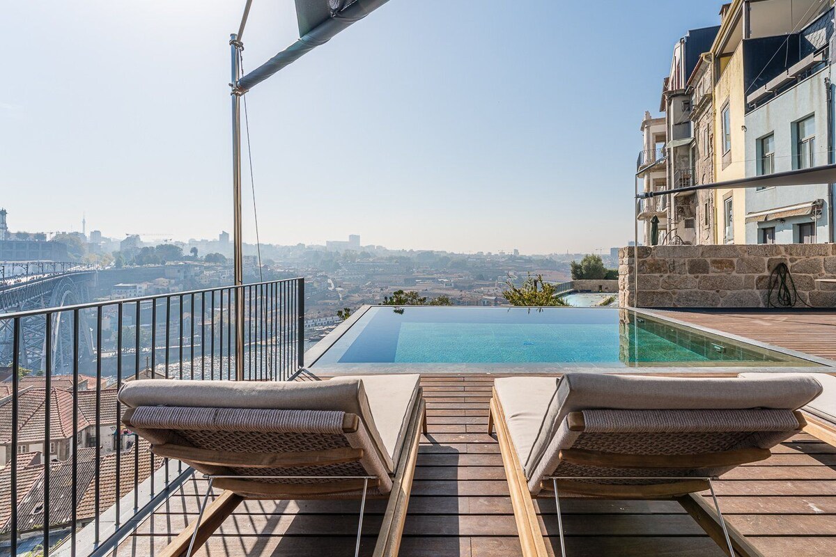 A rooftop infinity pool with two lounge chairs, overlooking a cityscape and a bridge.