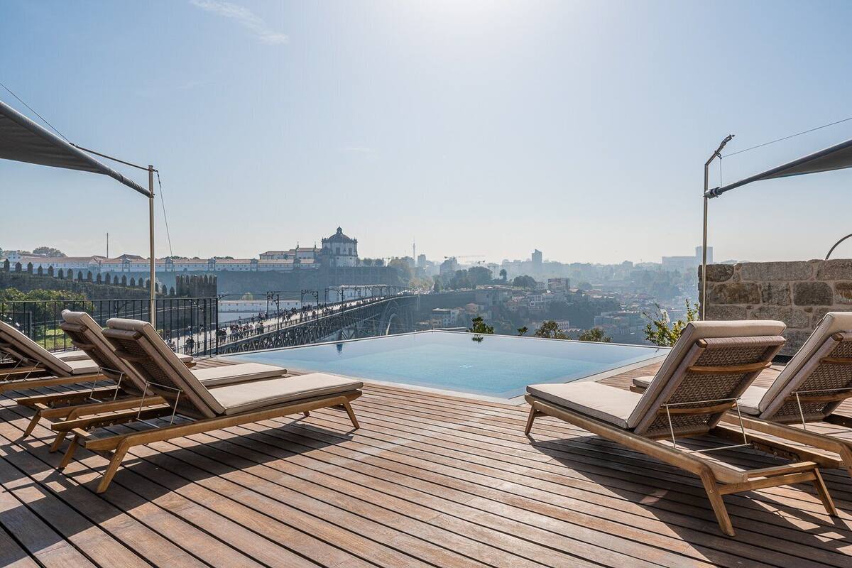 An infinity pool with lounge chairs, a view of a bridge, and a cityscape in the background.