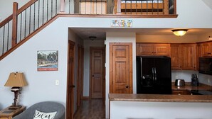 A spacious loft with a wooden staircase, a kitchen with wooden cabinets, a black refrigerator, and a cozy seating area with a gray sofa and a lamp. - The Good Life - Okoboji cabin in Bridges Bay (Arnolds Park)