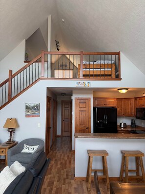 Un loft espacioso con escalera de madera, cocina con armarios de madera, refrigerador negro y una zona de estar acogedora con un sofá gris y una lámpara.