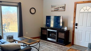 A living room with a ceiling fan, a television, a clock, and a door. - The Good Life - Okoboji cabin in Bridges Bay (Arnolds Park)