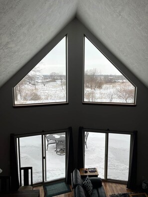 Ein Zimmer mit schrägen Wänden, großen Fenstern, einem schwarzen Ledersofa und Blick auf eine verschneite Landschaft.