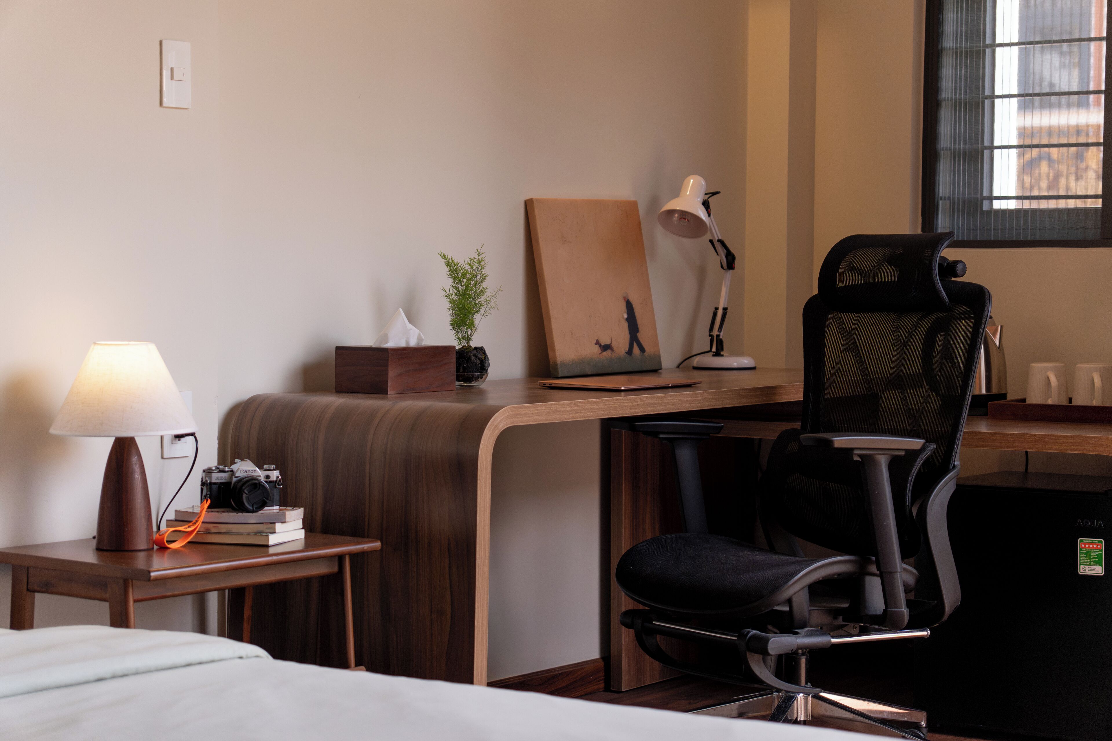 Een hotelkamer met een bed, een bureau met lamp, een stoel, een tafeltje met een camera en boeken, een potplant en een schilderij aan de muur.