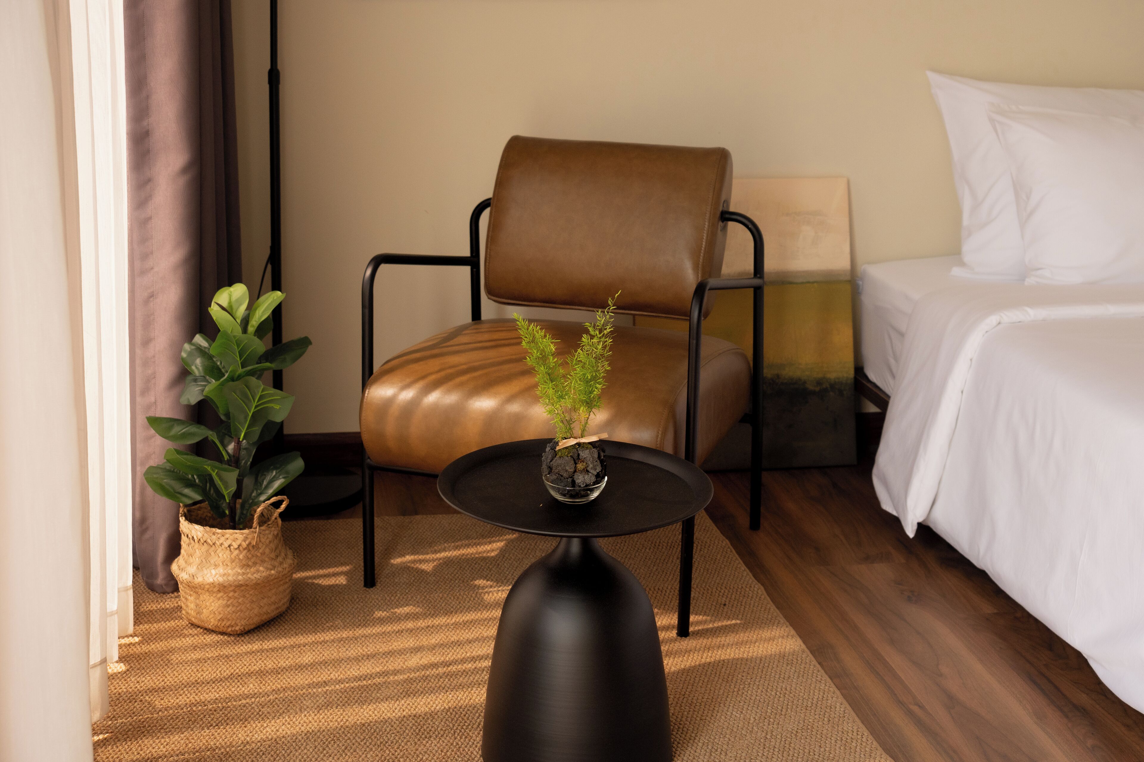 A hotel room with a bed, a chair, a small table with a plant, and a window with curtains.