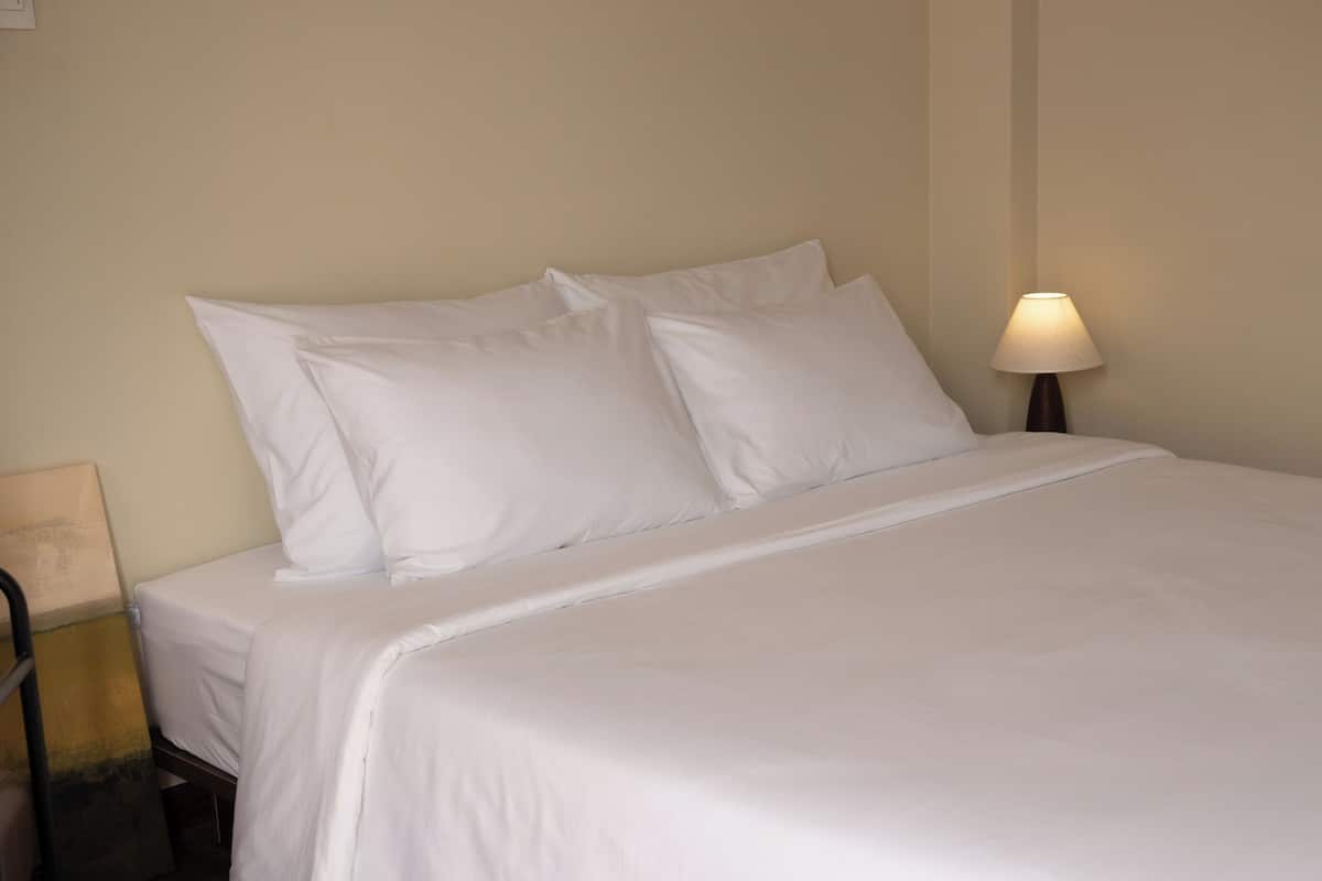 Een net opgemaakt bed met witte lakens, een bedlamp en een aan de muur bevestigd fotolijstje.