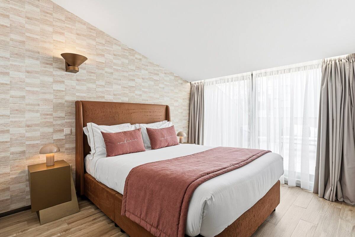 Una habitación de hotel moderna con pared de ladrillo, cabecero de madera, cama grande con ropa de cama blanca y rosa, y mesita de noche con lámpara.