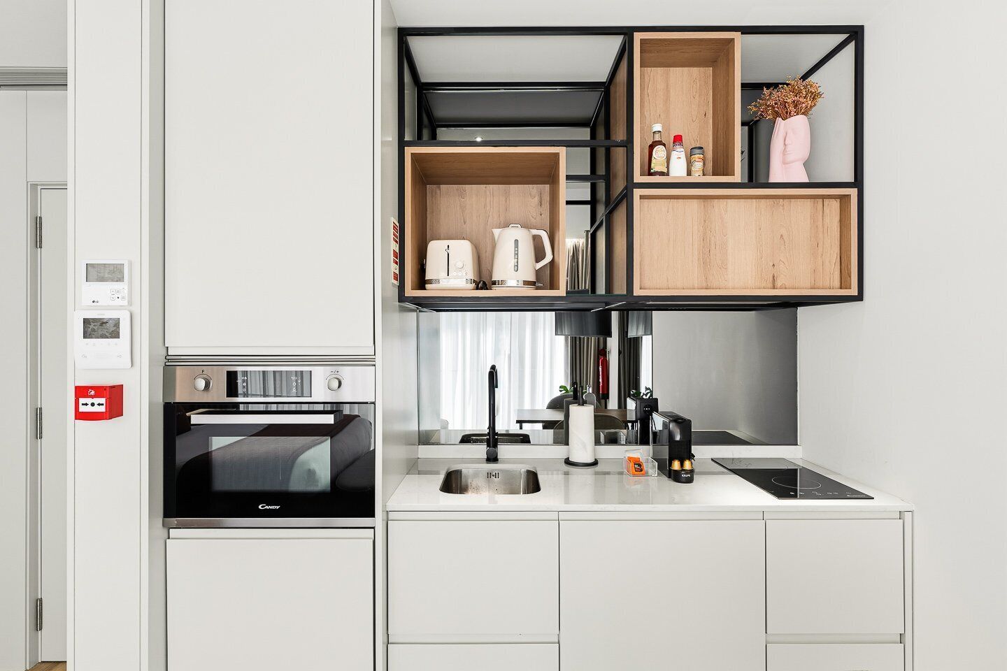 Una cocina compacta con horno integrado, cafetera y lavabo. Hay repisas de madera sobre la encimera que sostienen elementos esenciales de cocina.
