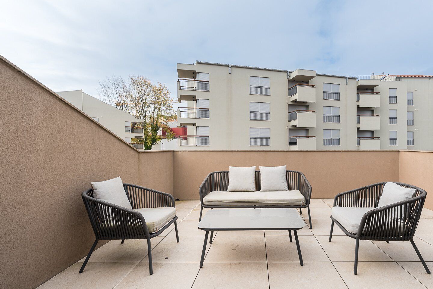Eine Terrasse im Freien mit Korbmöbeln, einem gefliesten Boden und Blick auf ein mehrstöckiges Gebäude im Hintergrund.