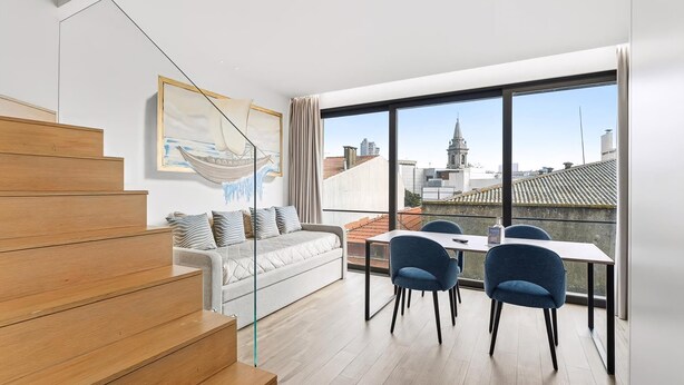 A modern apartment with a staircase, a sofa, a dining table with chairs, and large windows offering a city view. - Guestready - Alma Invicta (Porto)