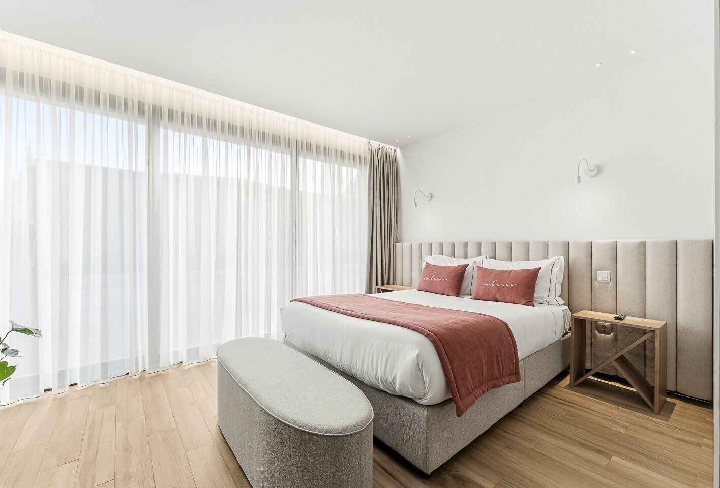 A modern hotel room with a large bed, a bedside table, a bench, and a large window with sheer curtains.