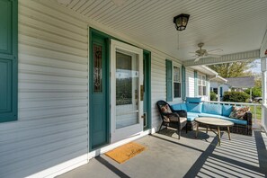 Eine Veranda mit weißer Fassade, grünem Zierstreifen, einem Korbsessel, einem Sessel und einem kleinen Tisch.