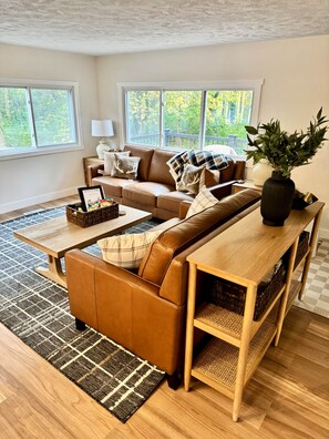 Ein Wohnzimmer mit braunem Ledersofa, einem Holztisch und Blick auf Bäume durch große Fenster.