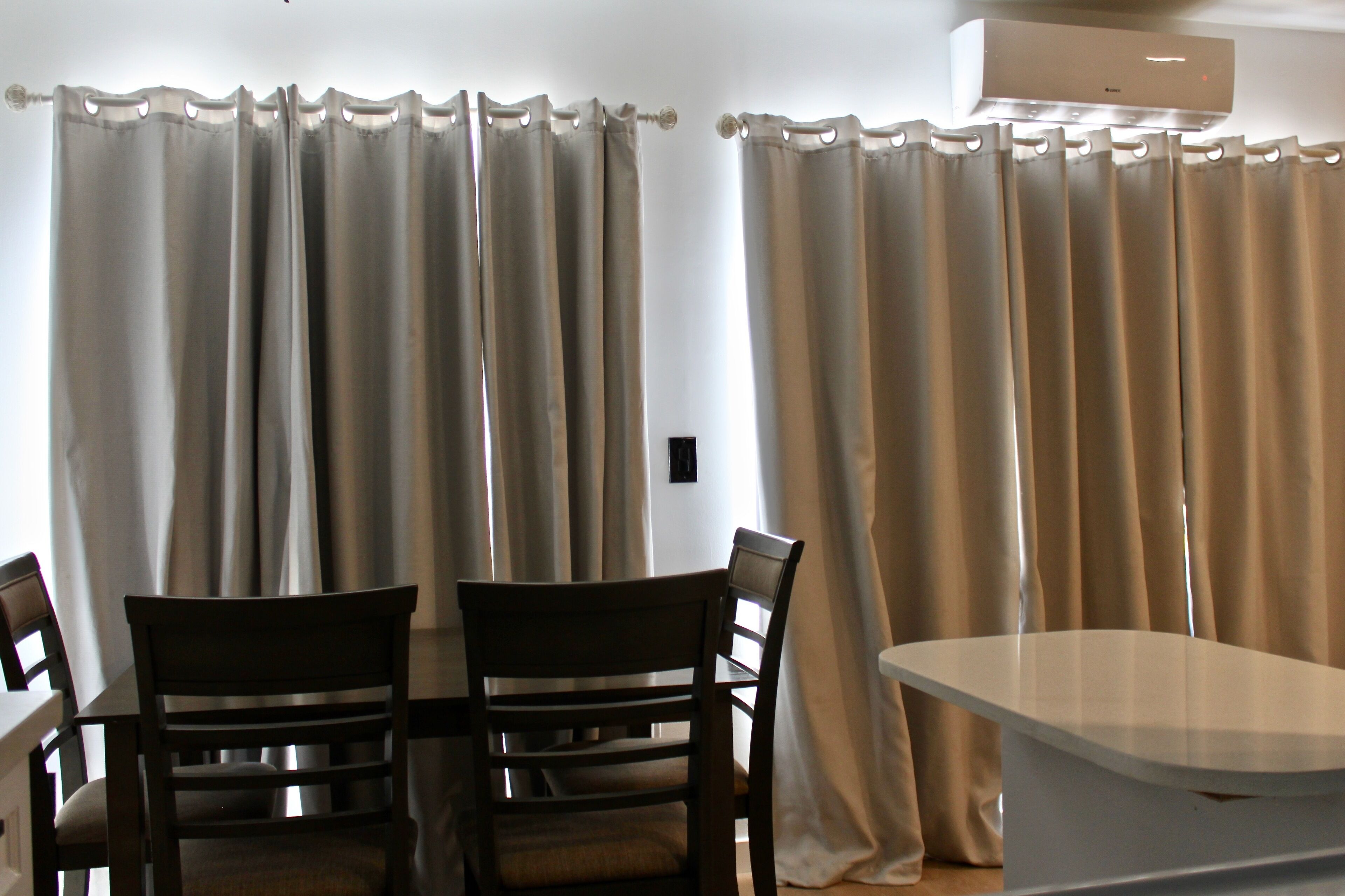 A dining area with a table and two chairs, a white ceiling-mounted air conditioner, and beige curtains.
