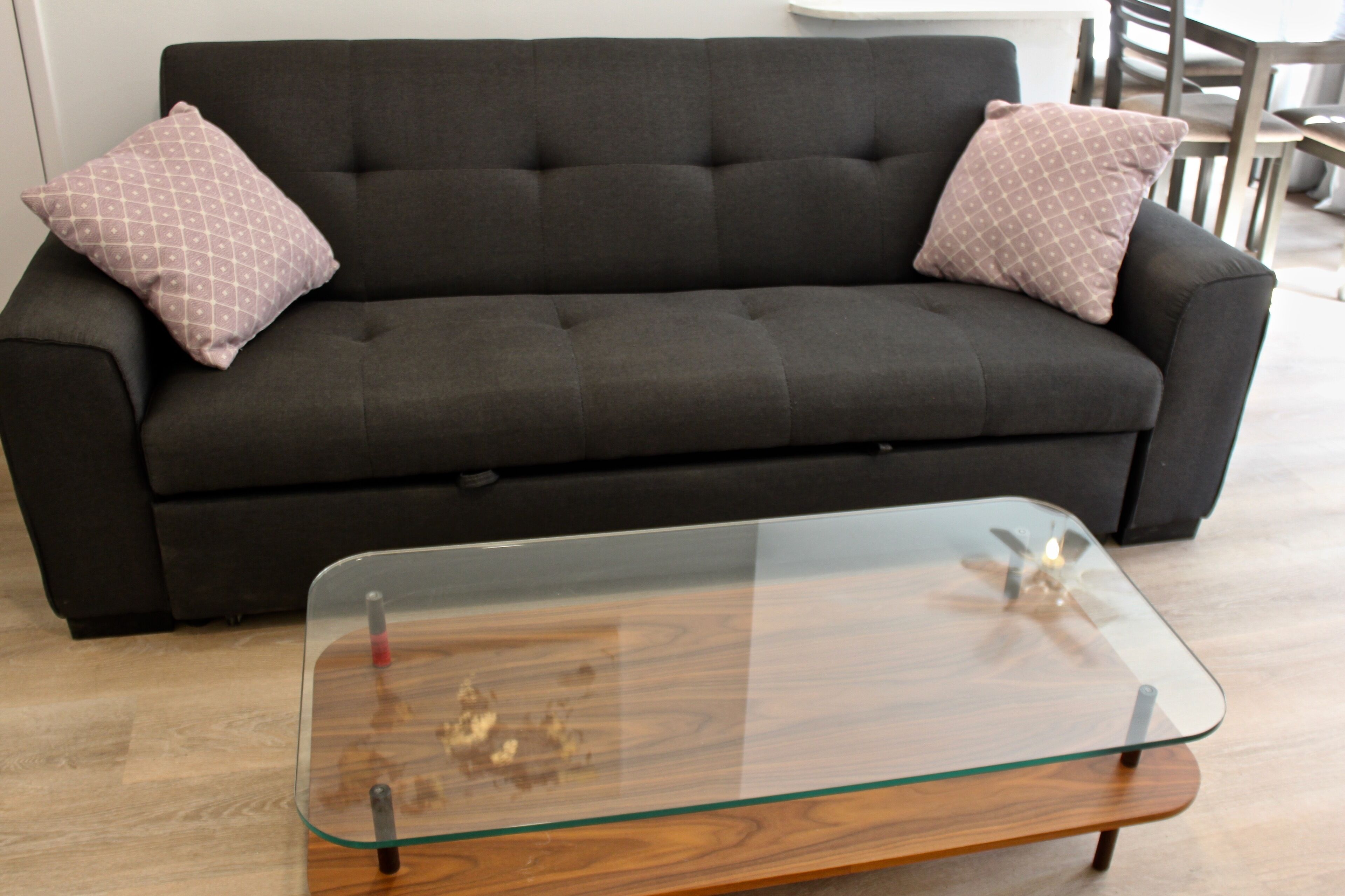 A dark grey sofa with two patterned cushions, a glass coffee table with wooden legs, and a wooden floor.