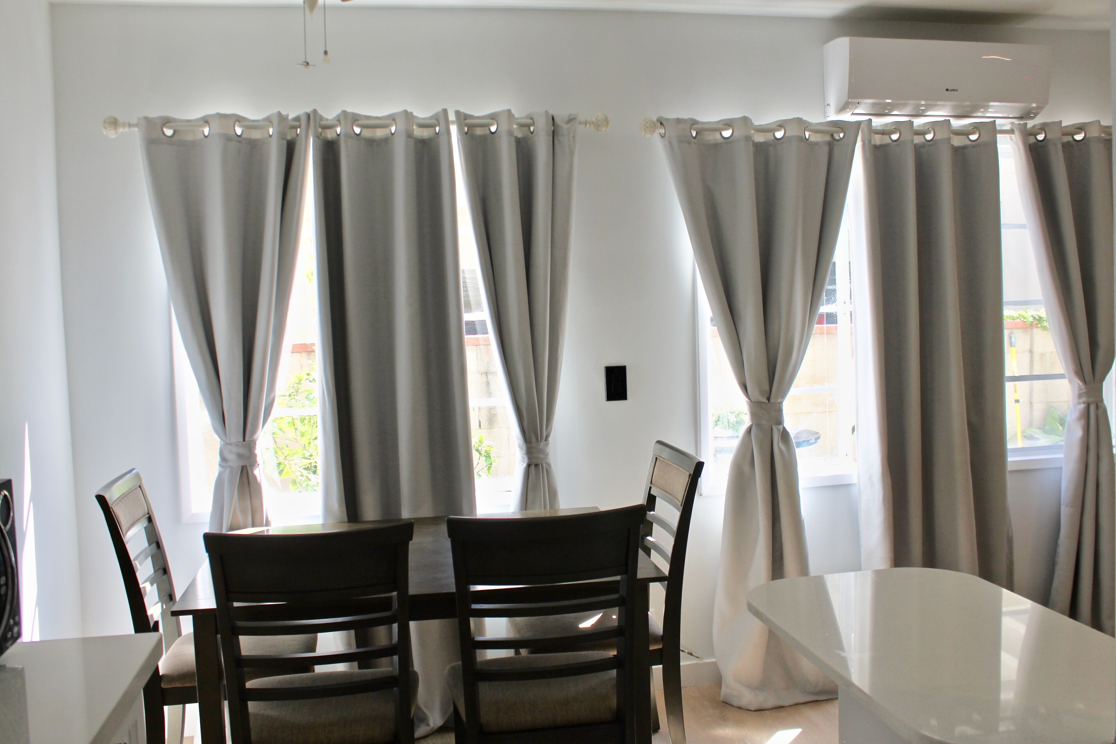 A dining area with a table and chairs, large windows with curtains, and an air conditioning unit.