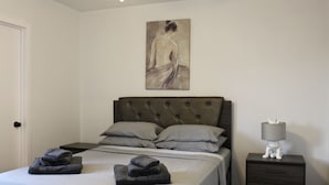 A neatly made bed with grey linens and pillows, a bedside table with a lamp, and a painting on the wall. - SMA Apartments (Santa Monica)