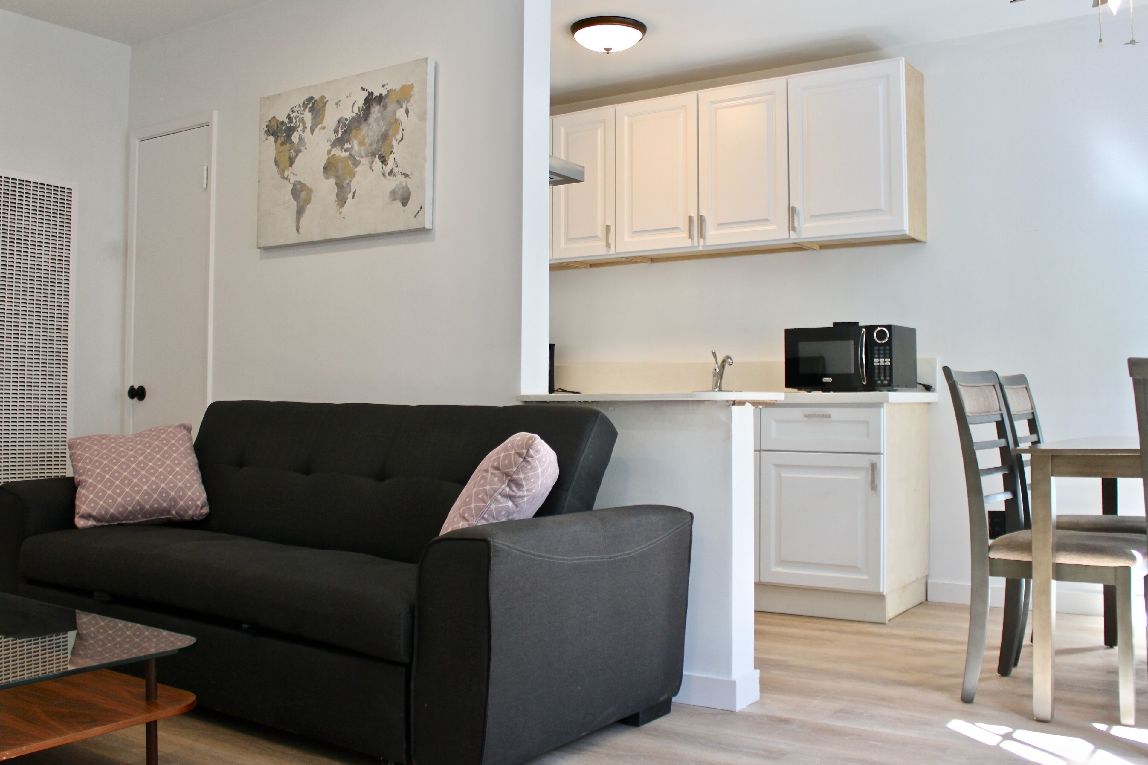 A modern living room with a black sofa, a wooden coffee table, and a world map on the wall. Adjacent is a kitchen with white cabinets and a microwave.