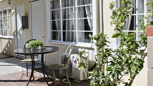 A patio with a table and chairs, a potted plant, and a window with curtains. - SMA Apartments (Santa Monica)