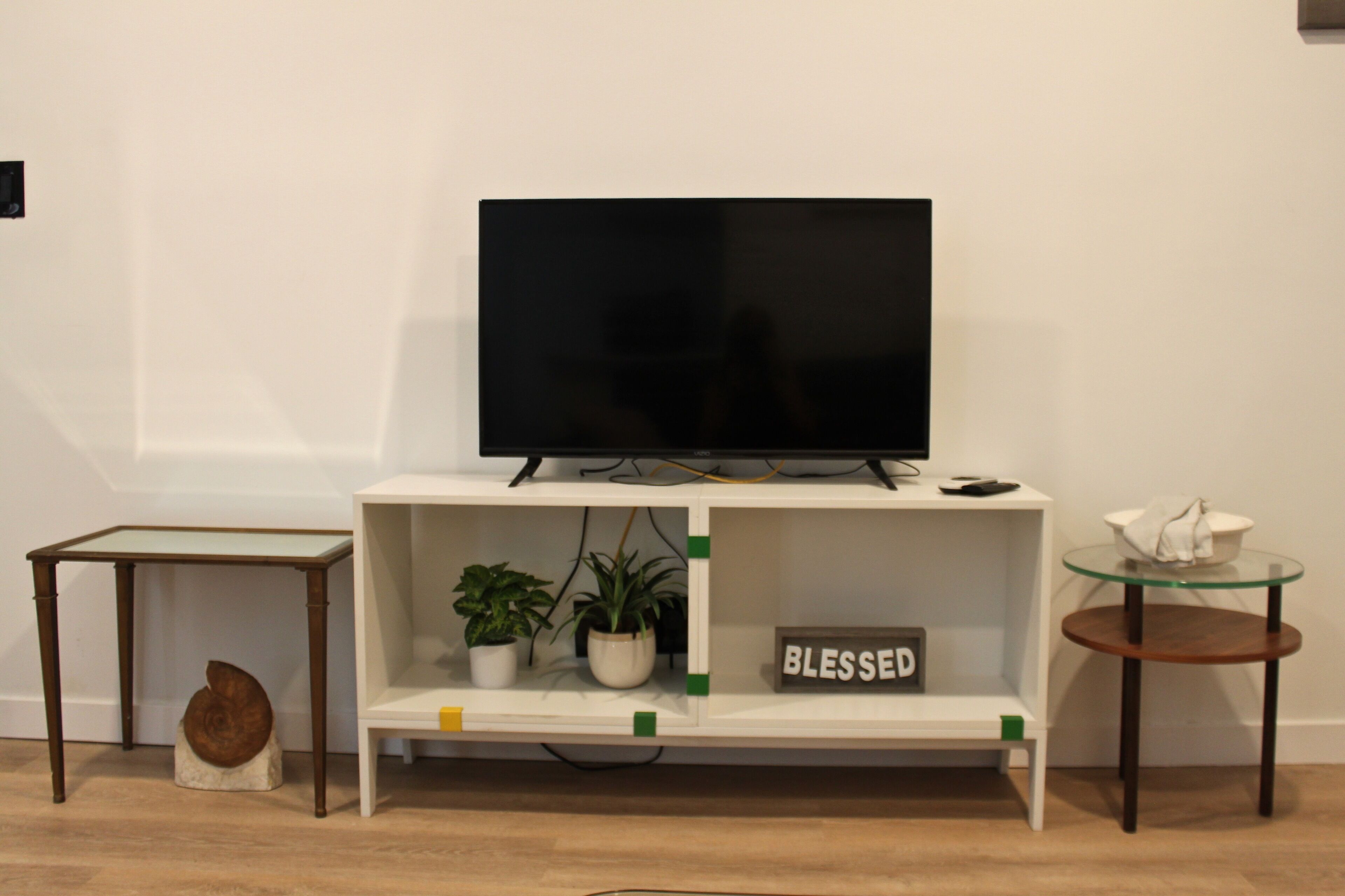 A modern living room with a flat-screen TV on a white stand, a small table with a potted plant, and a glass table with a bowl.