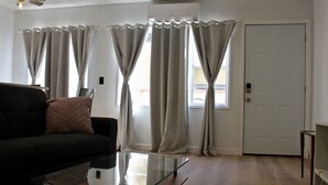 A living room with a glass coffee table, a black sofa with a patterned cushion, and light-colored curtains. - SMA Apartments (Santa Monica)