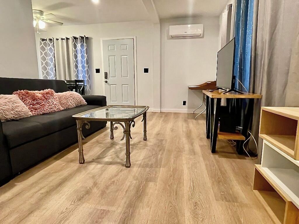 Un salón moderno con una mesa de centro de vidrio, un sofá negro con cojines rosa, un mueble de televisión de madera y una puerta al fondo.