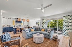 A modern living room with a sectional sofa, a round coffee table, wicker chairs, and a kitchen in the background. - Stunning Home w/ Pool, Slide & Spa & Rooftop Views (Sarasota)