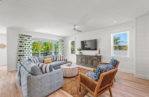 Ein geräumiges Wohnzimmer mit grauem Sofa, einem Korbsessel, einem runden Couchtisch und einem großen Fenster mit Jalousien.