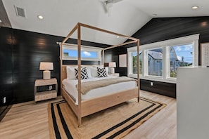 A four-poster bed in a modern bedroom with a dark wood finish, a wooden nightstand, a white lamp, and a view of a house through large windows. - Stunning Home w/ Pool, Slide & Spa & Rooftop Views! (Sarasota)