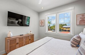 Quarto com cama, televisão na parede, janela com persianas e vista para o exterior.