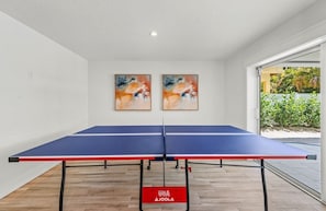 Uma mesa de pingue-pongue com superfície vermelha e azul, duas pinturas abstratas na parede e uma placa vermelha com texto branco no chão.