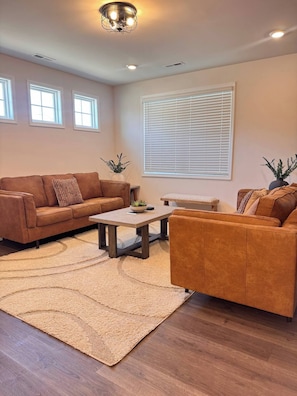 A living room with a brown sofa, a coffee table, a rug, and a large window with blinds. - Spacious Retreat w/ King Suite, 3 Baths + Yard (Graham)
