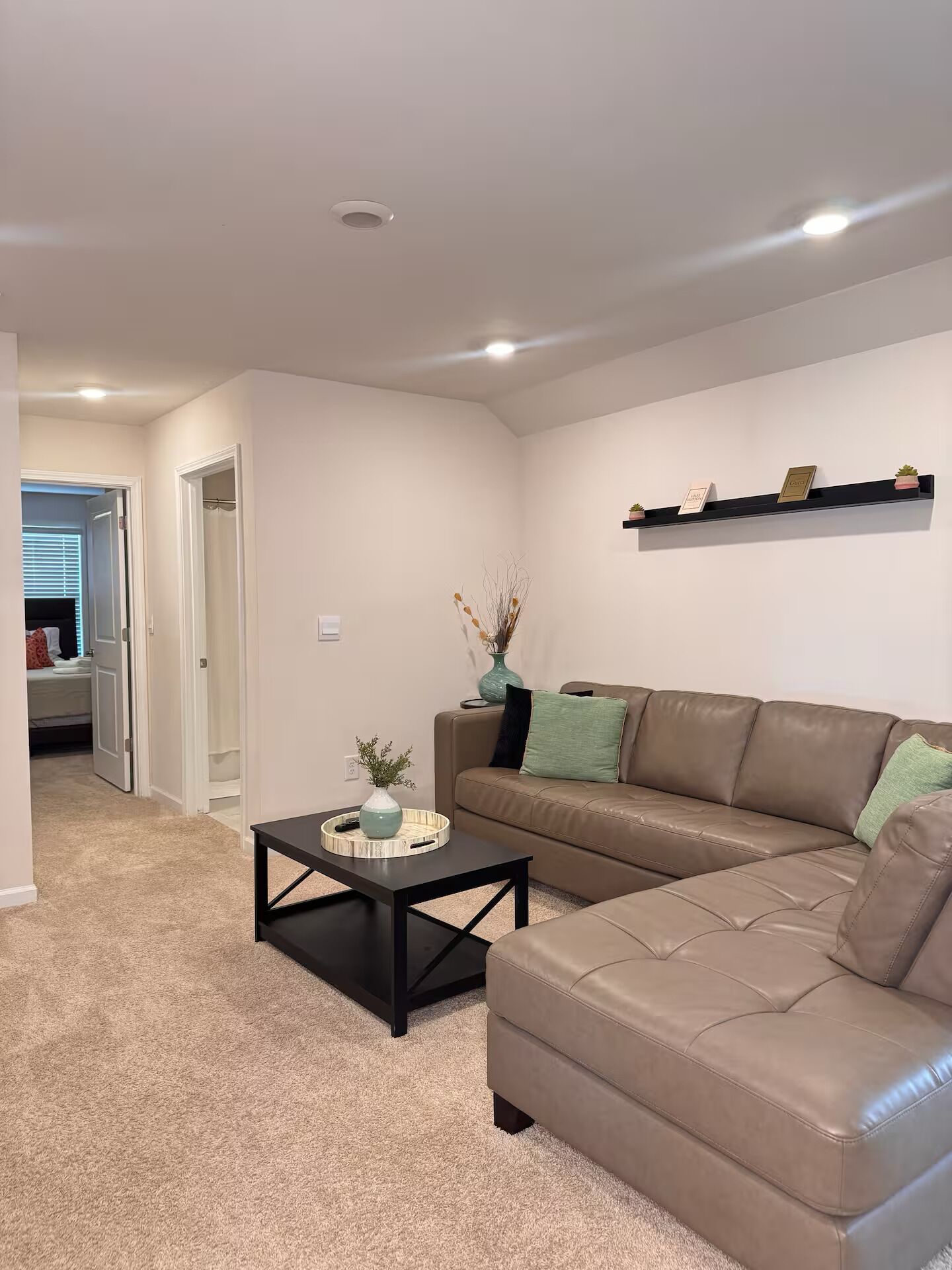 A living room with a brown leather sofa, a black coffee table, a vase with plants, and a shelf with decorative items.