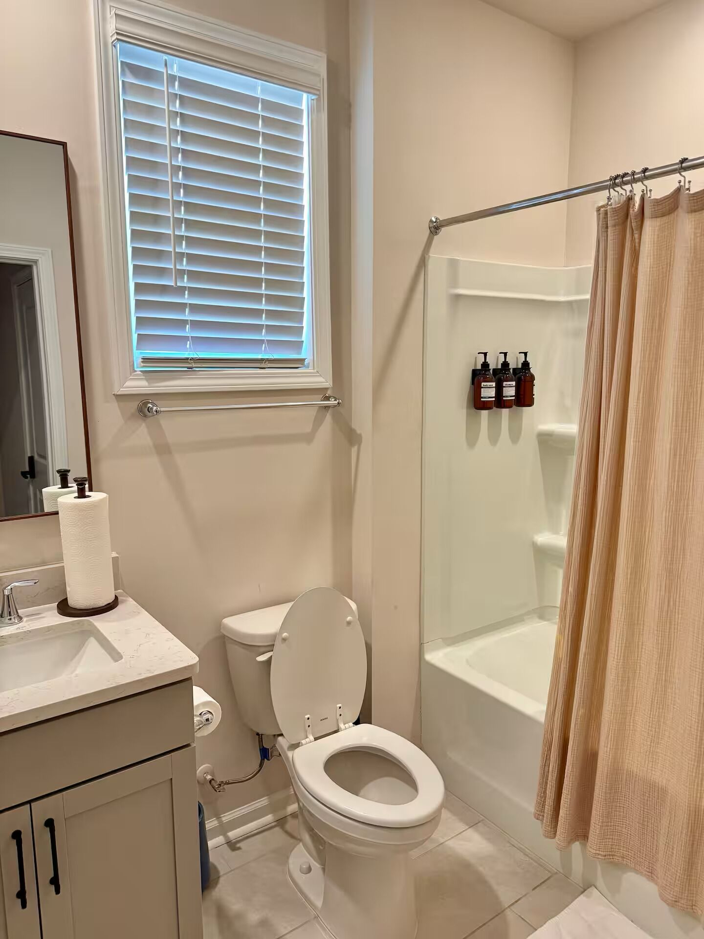 A bathroom with a toilet, sink, shower, and a window with blinds.