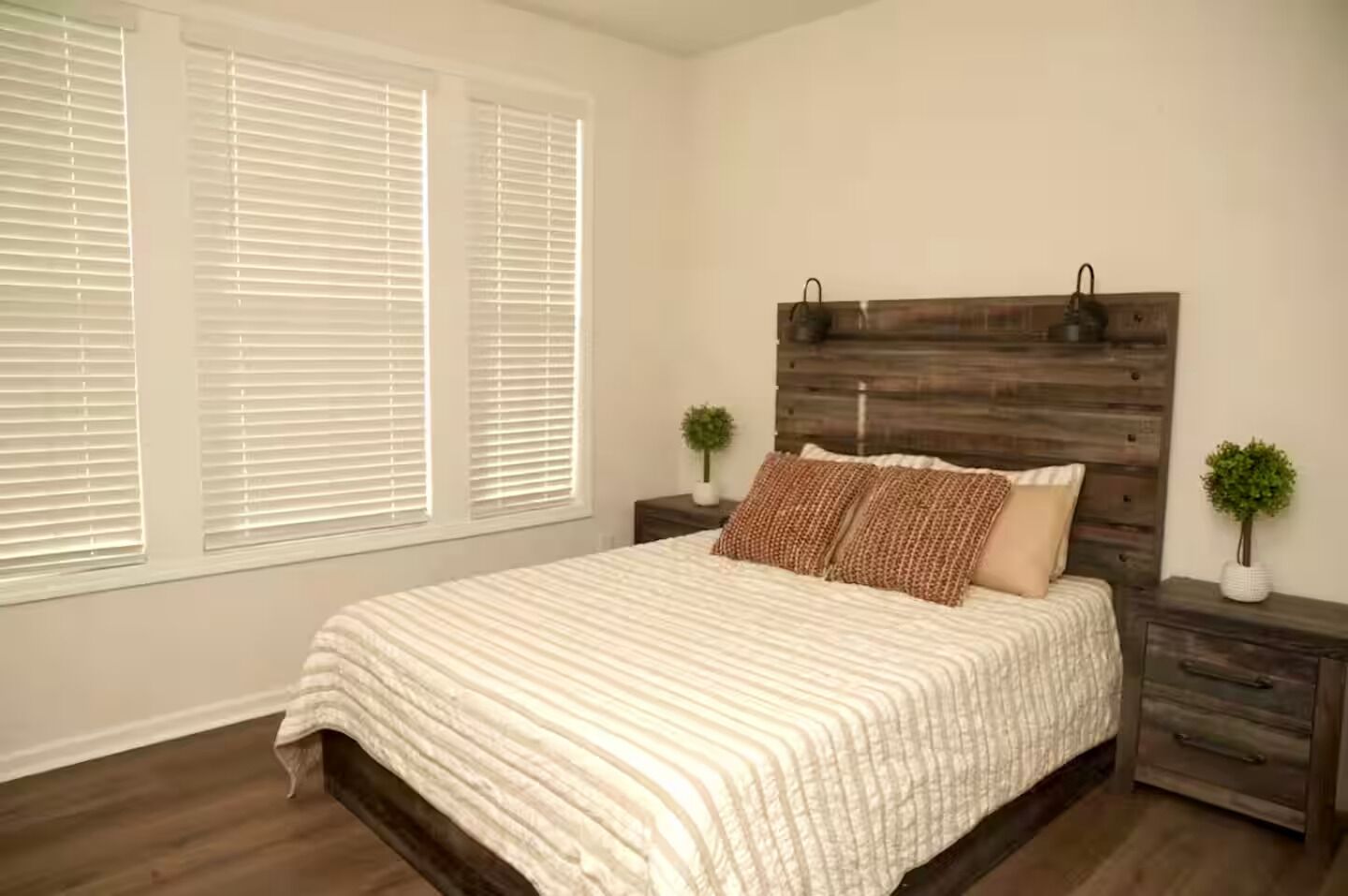 A bedroom with a bed, wooden headboard, two nightstands, a window with blinds, and a small plant on each nightstand.