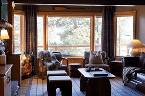 A cozy living room with a fireplace, a stone fireplace, a large window with curtains, a sofa, a coffee table, and a lamp. - Lakefront Cabin with Hot Tub and Dog Friendly (Bayfield)